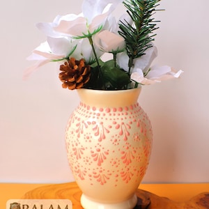 May include: A cream-colored ceramic vase with a pink floral pattern, holding white flowers, a pine cone, and a sprig of greenery. The vase has a rounded shape and a pedestal base. The logo "BALAM" is visible at the bottom.