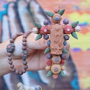 May include: A terracotta-colored cross pendant featuring a central relief of a figure, adorned with sculpted flowers in shades of red, blue, and beige, and green leaves. The cross is attached to a strand of brown spherical beads with flower accents.
