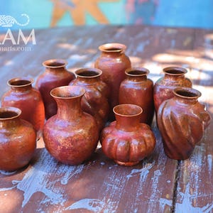 May include: A collection of ten small copper vases with varying shapes, including rounded, fluted, and twisted designs. Each vase features a warm, metallic reddish-brown patina with a textured finish and a narrow neck opening.