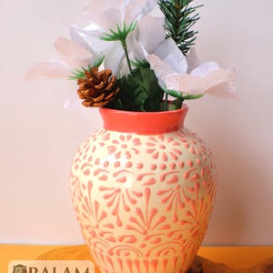 May include: A cream-colored ceramic vase with a coral rim and embossed pink floral patterns. The vase holds white artificial flowers, green leaves, a pine cone, and a sprig of evergreen. The logo "BALAM" is visible at the bottom.