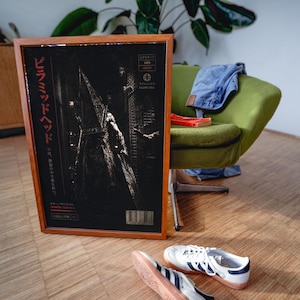 Puede incluir: P&oacute;ster enmarcado de Pyramid Head de Silent Hill, con texto japon&eacute;s y un c&oacute;digo de barras. La obra de arte es oscura con una figura con un abrigo largo. El marco es marr&oacute;n, y el p&oacute;ster se muestra cerca de una silla verde, vaqueros y zapatillas blancas.
