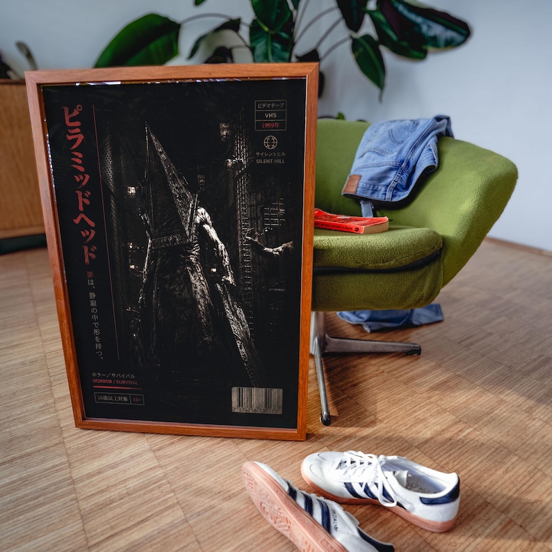 Puede incluir: P&oacute;ster enmarcado de Pyramid Head de Silent Hill, con texto japon&eacute;s y un c&oacute;digo de barras. La obra de arte es oscura con una figura con un abrigo largo. El marco es marr&oacute;n, y el p&oacute;ster se muestra cerca de una silla verde, vaqueros y zapatillas blancas.