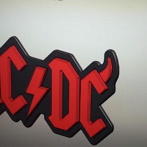 Puede incluir: Logotipo de AC/DC en negro y rojo con un rayo en el centro. El logotipo tiene un efecto 3D, con las letras y el rayo en rojo y delineados en negro. El fondo es gris claro.