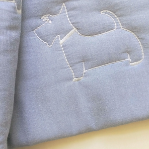 May include: A close-up of a pale blue fabric featuring a white embroidered Scottie dog. The dog is outlined in white thread, and the fabric has a subtle texture. This is likely part of a larger textile item.