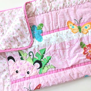 May include: Pink baby quilt with floral and butterfly appliqués. The quilt features a pink floral print on the border and a central panel with a leopard, butterflies, and flowers. The quilt is approximately 76 cm by 102 cm.