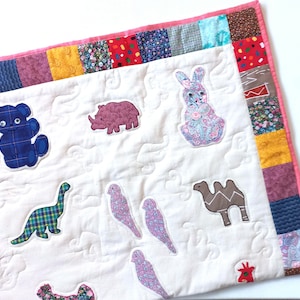May include: A handmade quilt with appliqued animal shapes: a blue teddy bear, a pink rhino, a floral bunny, a green dinosaur, three floral birds, and a brown camel. The quilt has a white background with quilted patterns and a colourful patchwork border.