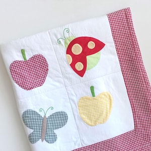 May include: A white quilted blanket featuring four square panels with fabric appliques of a red gingham apple, a red ladybird with beige spots, a grey gingham butterfly, and a yellow gingham apple, finished with a red gingham border trim.