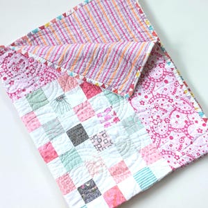 May include: A patchwork quilt in shades of pink, white, and pastel colours. The quilt has a paisley border and a striped pattern on the folded section. The quilt is made of soft fabric and is suitable for a baby or child. The quilt measures approximately 100 cm x 100 cm.
