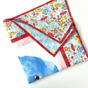 May include: A handmade quilt with a whimsical design. The quilt features a blue watercolor whale with a detailed eye. The edges are trimmed with a red border and a floral pattern. The quilt is made of soft, quilted fabric.