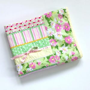 May include: A folded fabric quilt featuring a patchwork design with floral patterns in pink, white, and green. The quilt includes sections of striped and polka-dot fabric, accented with green rickrack trim, a yellow border, and a pink ribbon bow.