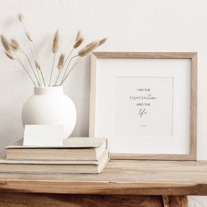 May include: A framed print with a white background and black text that reads "I am the resurrection and the life" with John 11:25 below the text. The print is in a light wood frame and is sitting on a wooden table with a stack of books and a white vase with dried flowers.