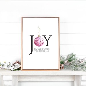 May include: A printable Christmas art print with a pink and white ornament and the word "JOY" in black text. The text below the ornament reads "Joy to the world, the Lord is come."