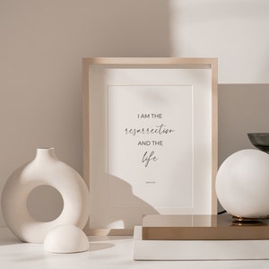 May include: A framed print with a white background and black text that reads "I am the resurrection and the life" with "John 11:25" below the text. The print is sitting on a wooden shelf with a white vase and a white globe lamp.