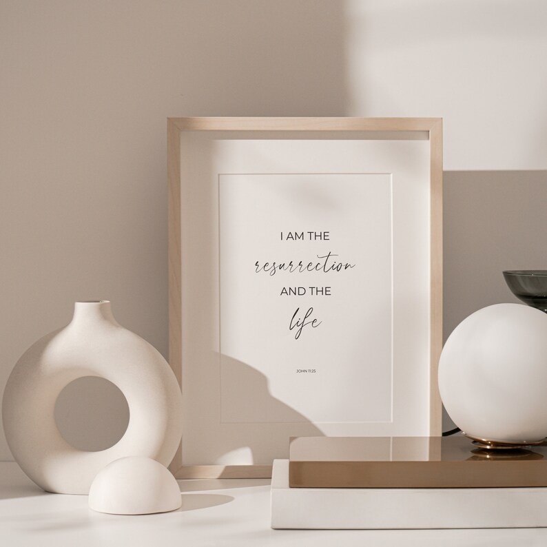 May include: A framed print with a white background and black text that reads "I am the resurrection and the life" with "John 11:25" below the text. The print is sitting on a wooden shelf with a white vase and a white globe lamp.