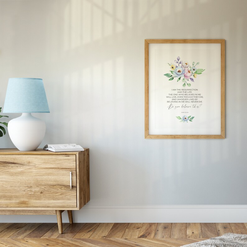 May include: A framed print with a light wood frame. The print features a floral watercolor design in pastel colors above text that reads "I AM THE RESURRECTION AND THE LIFE... Do you believe this?" The print hangs on a light gray wall.