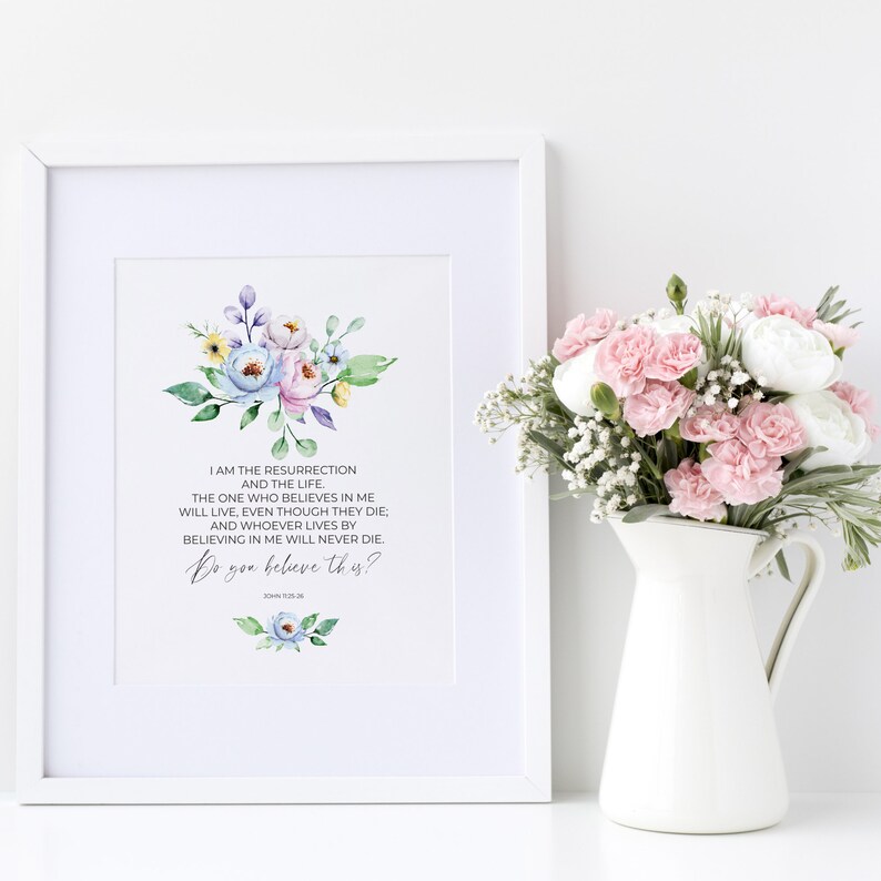 May include: A framed print with a floral watercolor design and the text "I am the resurrection and the life... Do you believe this?" A white pitcher holds a bouquet of pink and white flowers. The print is in a white frame.