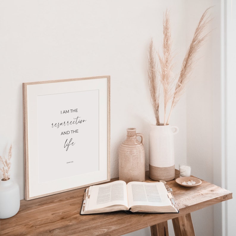 May include: A framed print with a white background and black text that reads "I am the resurrection and the life" with the scripture reference "John 11:25" below. The print is on a wooden table with a Bible, a vase with dried pampas grass, and a ceramic jug.