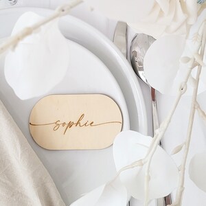 May include: A wooden place card with the name "Sophie" engraved in cursive script. The place card is oval shaped and is sitting on a white plate.