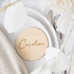 May include: A round wooden place card with the name "Caroline" engraved in a cursive font. The place card is on a white plate with a white napkin and white flowers.