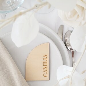 May include: A wooden place card with a rounded edge and the name "CAMILIA" engraved in gold lettering. The place card is on a white plate with a white napkin and white flowers.