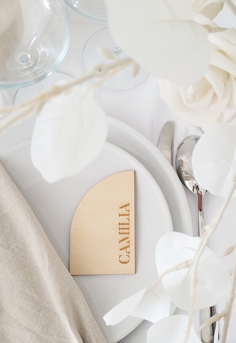 May include: A wooden place card with a rounded edge and the name "CAMILIA" engraved in gold lettering. The place card is on a white plate with a white napkin and white flowers.