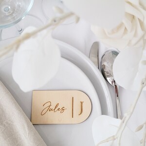 May include: A wooden place card with the name "Jules" and the initial "J" engraved on it. The place card is on a white table setting with a white napkin and a silver fork.