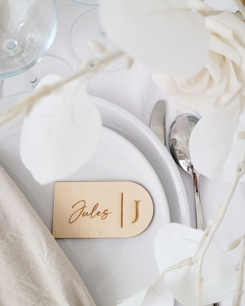 May include: A wooden place card with the name "Jules" and the initial "J" engraved on it. The place card is on a white table setting with a white napkin and a silver fork.