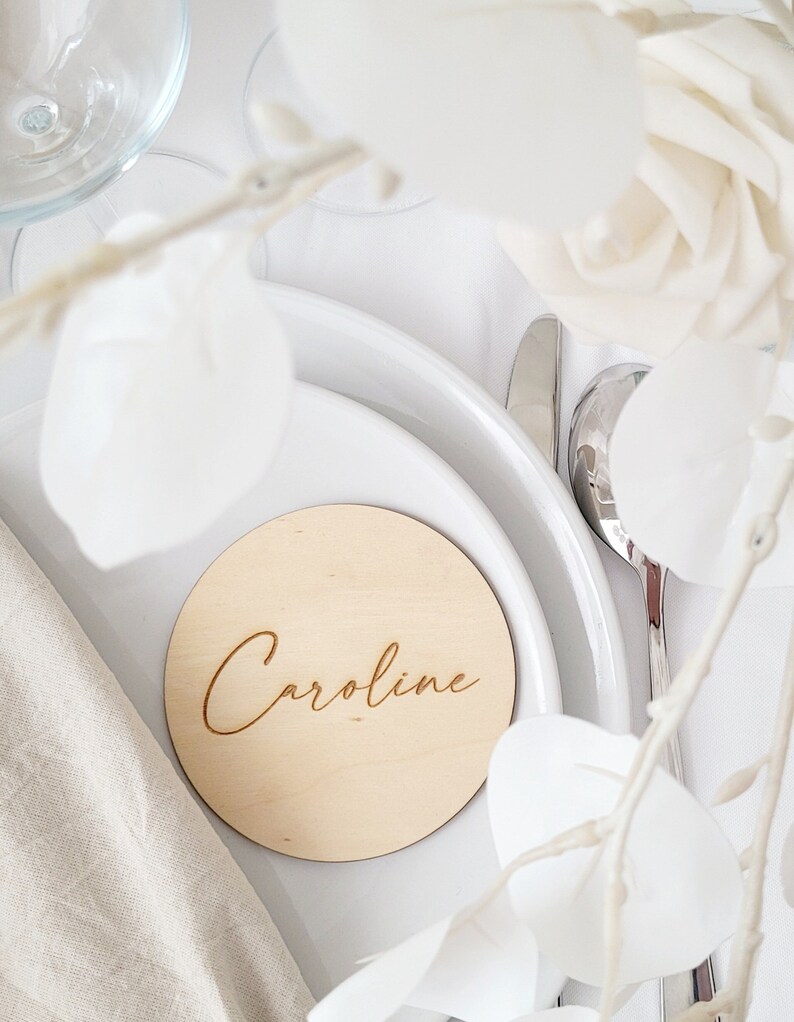 May include: A round wooden place card with the name "Caroline" engraved in a cursive font. The place card is on a white plate with a white napkin and white flowers.