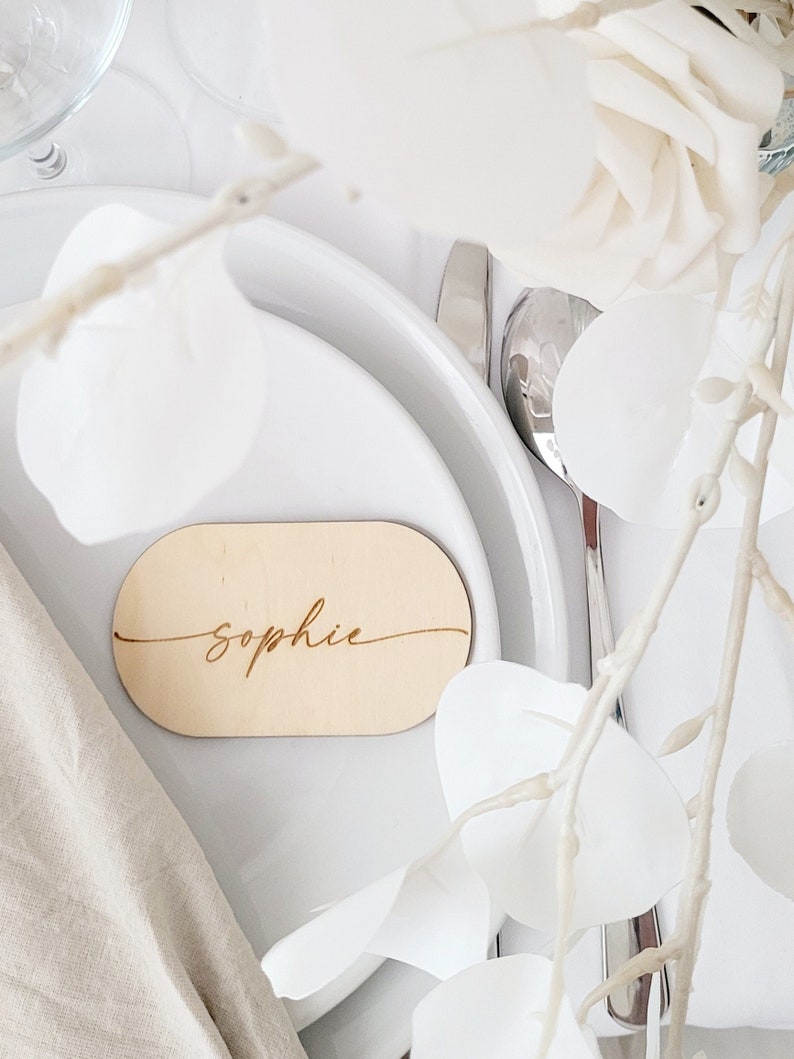 May include: A wooden place card with the name "Sophie" engraved in cursive script. The place card is oval shaped and is sitting on a white plate.