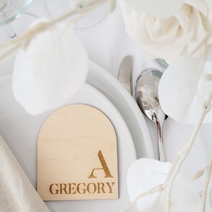 May include: A wooden place card with a rounded arch top. The place card is engraved with the letter "A" and the name "GREGORY".