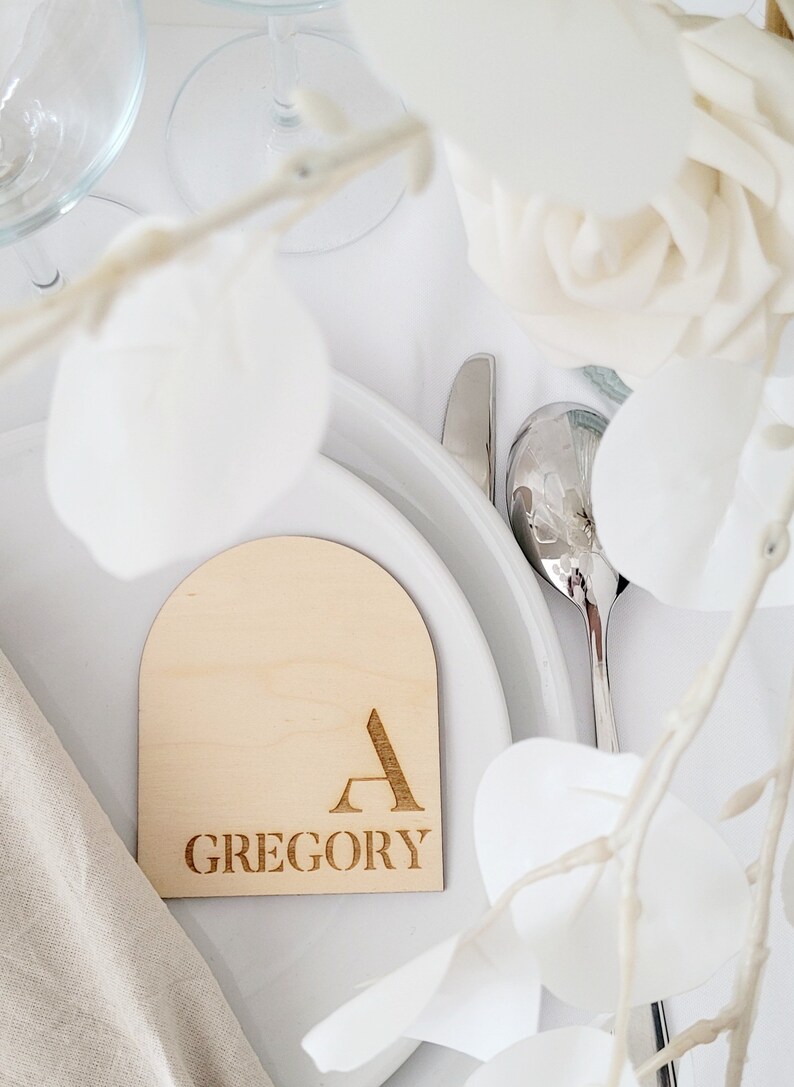 May include: A wooden place card with a rounded arch top. The place card is engraved with the letter "A" and the name "GREGORY".