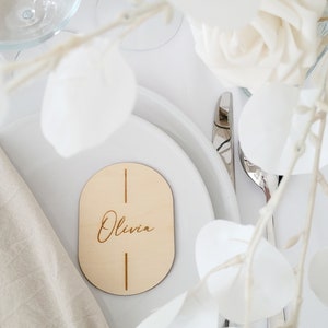 May include: A wooden oval place card with the name "Olivia" engraved in cursive script. The card is on a white plate with a white napkin and a silver fork.