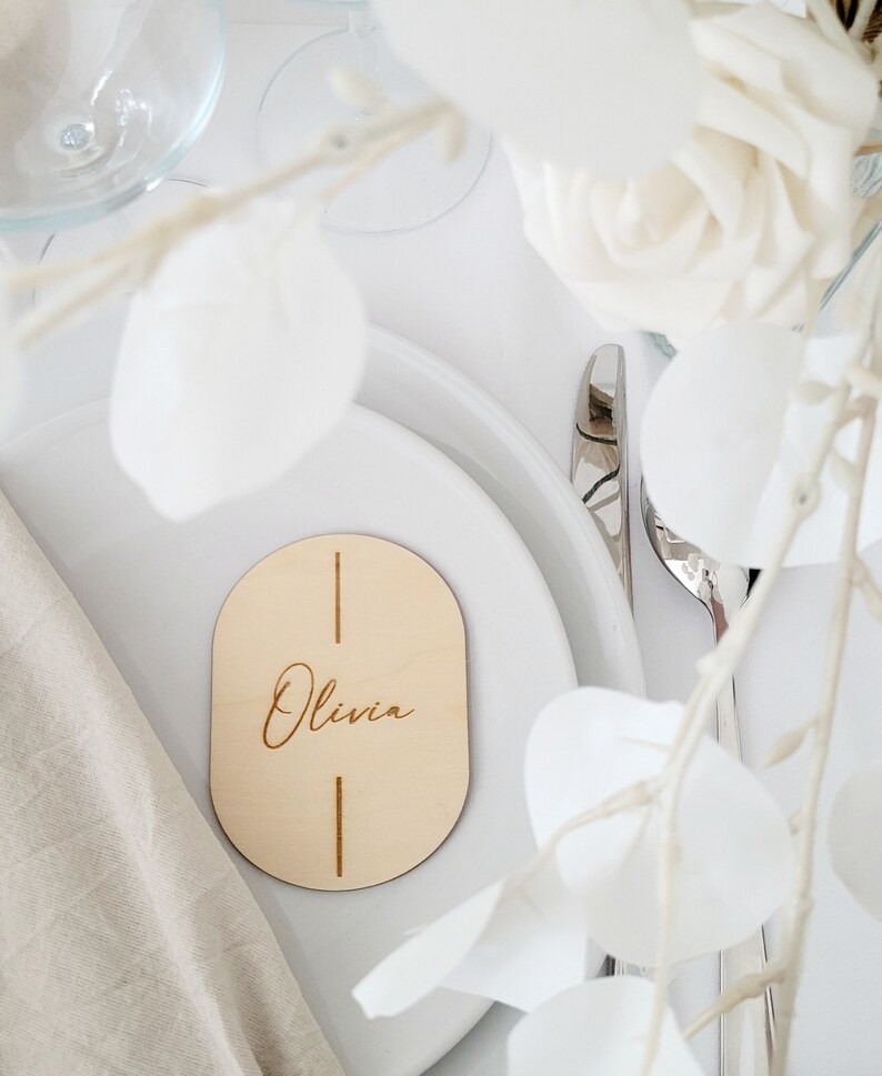 May include: A wooden oval place card with the name "Olivia" engraved in cursive script. The card is on a white plate with a white napkin and a silver fork.