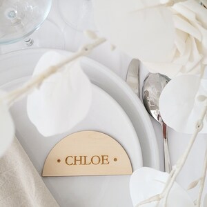 May include: A wooden place card with a rounded top and the name "CHLOE" engraved in the center. The place card is on a white plate with a white napkin and a silver fork.