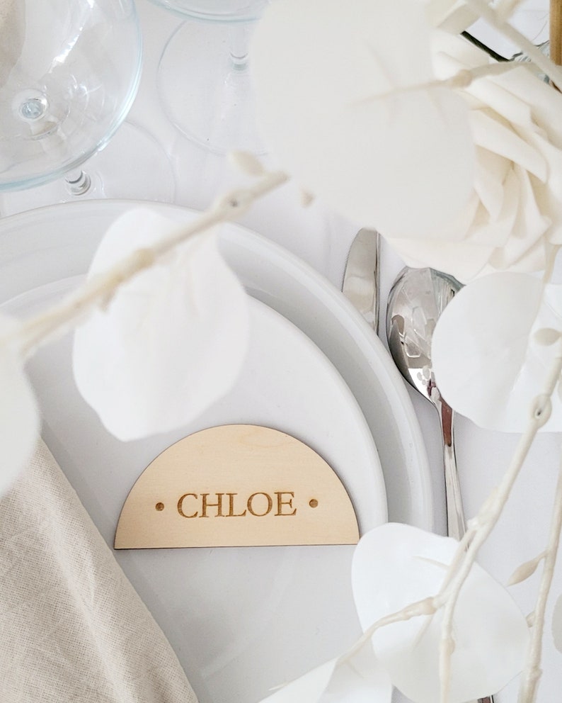 May include: A wooden place card with a rounded top and the name "CHLOE" engraved in the center. The place card is on a white plate with a white napkin and a silver fork.