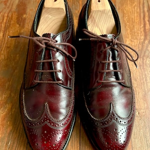 May include: A pair of burgundy leather dress shoes with brogue detailing and brown laces. The shoes have wooden shoe trees inside. The shoes are displayed on a wooden surface.