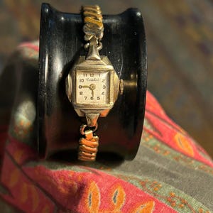 May include: A vintage gold-toned wristwatch with a square face and a cream-colored dial. The watch features the brand name "Enicar" and gold-toned, flexible bands. The watch is displayed on a black, curved object.