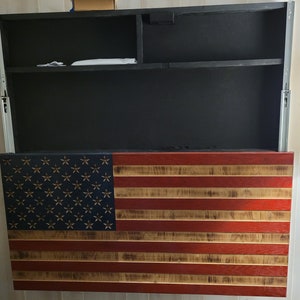 Puede incluir: Una bandera americana de madera con colores rojo, blanco y azul. La bandera está montada en una pared y tiene un compartimento oculto detrás. El compartimento está abierto y revela dos estantes.