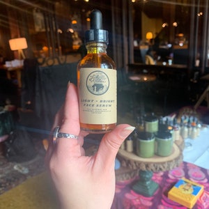 May include: A small amber glass bottle with a dropper top. The bottle is labeled "Lady Bird Botanicals" and "Light + Bright Face Serum". The bottle is held in a hand with a silver ring.
