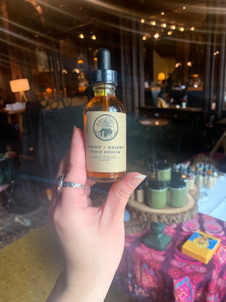 May include: A small amber glass bottle with a dropper top. The bottle is labeled "Lady Bird Botanicals" and "Light + Bright Face Serum". The bottle is held in a hand with a silver ring.