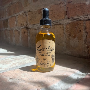 May include: A glass bottle with a dropper top filled with a light yellow liquid. The bottle has a brown paper label with the text "Light + Bright Face Oil by Lady H" in black.