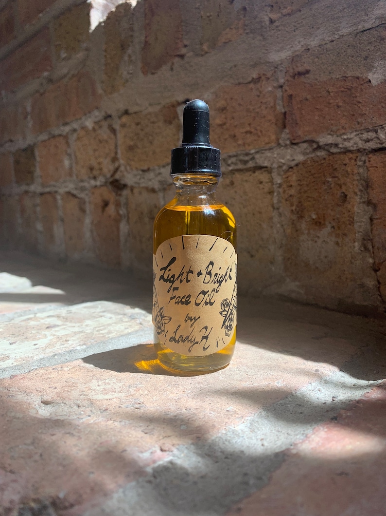 May include: A glass bottle with a dropper top filled with a light yellow liquid. The bottle has a brown paper label with the text "Light + Bright Face Oil by Lady H" in black.