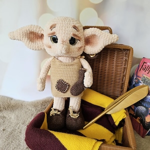 May include: A handmade crochet doll with large ears, wearing a tan tunic and brown shoes, holding a small brown object. The doll is in a wicker basket with a yellow and burgundy scarf and a golden feather pen.