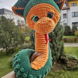 May include: A handmade crochet snake toy in shades of green and orange. The snake has large, expressive eyes, a red tongue, and a textured body. The toy is held in a person's hand, showcasing its size and detail.