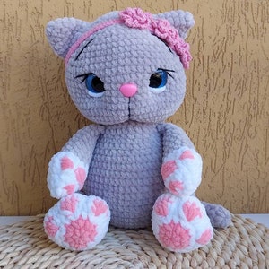 May include: A grey crocheted cat toy with pink and white paw details and a pink flower headband. The cat is sitting on a woven straw mat.