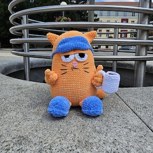May include: A crocheted orange cat toy wearing a blue eye mask and holding a white coffee mug. The cat is sitting on a gray concrete surface.