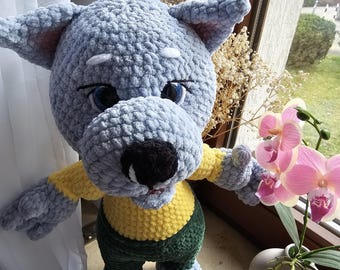 Puede incluir: Un juguete de peluche de lobo de crochet que lleva una camisa amarilla y verde. El lobo tiene ojos azules y una nariz negra. El juguete está sentado en un alféizar de ventana con un jarrón de orquídeas rosas en el fondo.