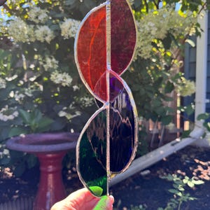 May include: A stained glass leaf-shaped suncatcher with a silver-colored metal frame. The suncatcher is divided into four sections, with colors ranging from red to purple, and green to blue. The suncatcher is hanging outside.
