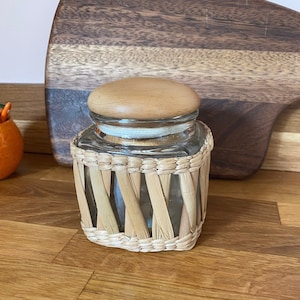 May include: A clear glass jar with a wooden lid and a woven wicker basket around the middle. The jar is sitting on a wooden surface.