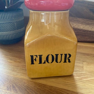 Może przedstawiać: Ceramiczny pojemnik na mąkę z czerwoną pokrywką. Pojemnik jest żółty i ma napis "FLOUR" nadrukowany czarnymi literami.